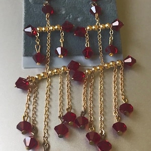 May include: Dangling earrings with a gold-tone metal design. Each earring features a series of faceted, deep red beads and delicate gold chains. The earrings are displayed on a gray card with the text "ACCESSORY PLACE".