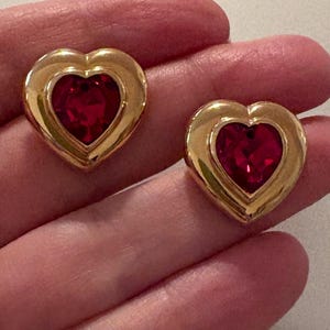 May include: A pair of heart-shaped earrings featuring a polished gold-tone metal frame surrounding a faceted, deep red gemstone center. Each earring displays a smooth, rounded outer edge and a pointed base, creating a classic heart silhouette.