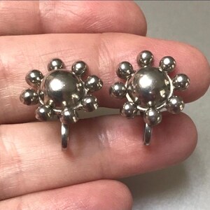 May include: A pair of silver-coloured clip-on earrings. Each earring has a central sphere surrounded by smaller spheres, forming a floral or sunburst pattern. The earrings are held by a person's hand.