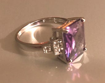 Puede incluir: Un anillo plateado con una gran piedra preciosa rectangular púrpura. El anillo tiene una banda con pequeñas piedras de acento en el lateral de la piedra principal. El anillo está sobre una superficie clara.