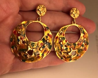 Puede incluir: Un par de pendientes de tono dorado con un diseño circular. Los pendientes están decorados con un patrón de esmalte colorido en tonos de rojo, verde, azul y amarillo.