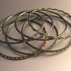 May include: A collection of gold-toned bangles. Some bangles have a smooth, solid design, while others feature intricate patterns of small, sparkling embellishments. The bangles are stacked together, creating a layered effect.