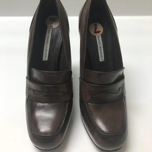 May include: A pair of brown leather heeled loafers. The shoes have a rounded toe, a strap across the vamp, and a stacked heel. The brand name "VERA WANG LAVENDER" is printed on the inner sole.