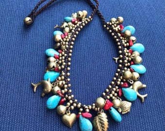 Puede incluir: Collar artesanal con cuentas turquesas en forma de lágrima, pequeñas campanas doradas y varios dijes, incluyendo delfines, corazones y hojas. El collar está ensartado en un cordón marrón oscuro.