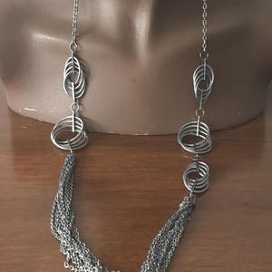 以下が含まれることがあります： 楕円形と円形の金属要素が連なったシルバーカラーのネックレス。ネックレスは、下方に連なる複数のチェーンストランドが特徴です。ジュエリーはマネキンに展示されています。