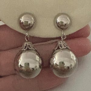 May include: A pair of silver-toned drop earrings featuring a smooth, spherical ball suspended from a smaller, matching silver-toned dome stud. Each large sphere is attached to the stud by a small metal link and features a decorative filigree cap.