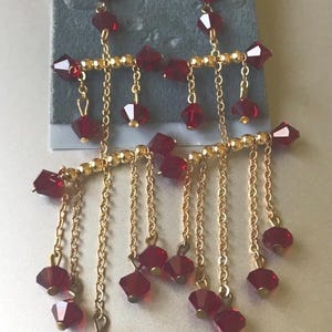 May include: Dangling earrings with a gold-tone metal design. The earrings feature faceted, deep red beads and chains. The top of the earrings are attached to a gray card with the text "ACCESSORY PLACE".