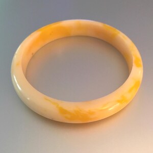 May include: A smooth, circular bangle bracelet featuring a marbled pattern of creamy off-white and vibrant yellow tones. The bracelet has a polished, glossy finish and a rounded, continuous band design that maintains a consistent thickness throughout.