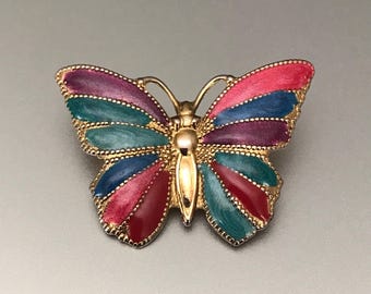 Puede incluir: Broche de mariposa dorada con alas de esmalte colorido. Las alas presentan secciones rojas, rosas, azules, turquesas y moradas. El broche tiene un borde y antenas doradas texturizadas.
