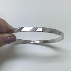 May include: A silver bangle bracelet with a textured, diagonal stripe design. The bracelet is round and has a polished finish. The bangle is held by a hand with a painted fingernail.