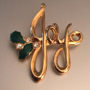 May include: A gold-coloured brooch spelling "Joy" in a cursive font. The brooch features a holly leaf design with green enamel and three clear, round rhinestones. A festive accessory.