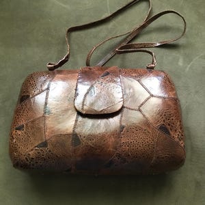 May include: A brown handbag constructed from multiple stitched leather patches with varying textures and shades. The bag features a rectangular shape with rounded corners, a flap closure, and a thin, adjustable shoulder strap attached at the top.