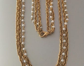 Puede incluir: Un collar dorado con múltiples hilos. El collar presenta una combinación de eslabones de cadena dorados y hilos de pequeñas perlas blancas de imitación. El cierre también es dorado. El collar se muestra sobre un fondo claro.