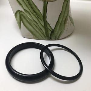 May include: Two black bangles, one thick and one thin, are displayed on a white surface. A decorative vase with a tulip design in red, green, and white is in the background.