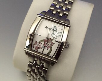 Puede incluir: Un reloj Armitron plateado con una esfera rectangular. La esfera blanca del reloj presenta detalles florales rosas y la palabra "Now". El reloj tiene una correa de eslabones plateada.