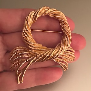 Puede incluir: Broche dorado con un diseño texturizado en forma de cuerda. El broche presenta una parte superior en forma de lazo y una parte inferior anudada y fluida, asegurada con un alfiler. El diseño general es elegante y de inspiración vintage.