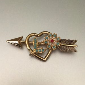 May include: Gold-toned brooch featuring a heart pierced by an arrow. The heart is adorned with a flower and turquoise-colored accents. The arrow has a detailed feather design, creating a vintage-inspired accessory.