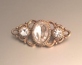 May include: A gold-toned metal brooch featuring an elongated oval clear crystal at the center, flanked by two smaller round clear crystals. The metal frame displays intricate filigree scrollwork and beaded detailing surrounding each stone.
