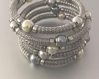 Puede incluir: Una pulsera de espiral de múltiples hilos con tubos de malla en tono plateado intercalados con cuentas redondas en tonos crema, blanco y gris metálico. El diseño flexible se envuelve sobre sí mismo, creando una apariencia en capas con acentos alternos de metal y cuentas tipo perla.