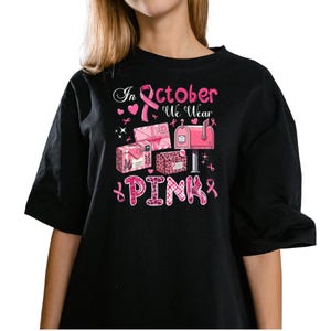 May include: Black t-shirt with a graphic design featuring pink mailboxes, gift boxes, and ribbons. The text "In October We Wear Pink" is displayed in pink and white, promoting breast cancer awareness.