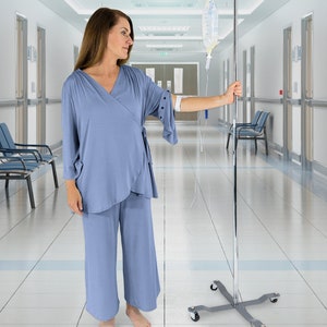 Puede incluir: Una mujer con una bata de hospital azul claro se encuentra en un pasillo de hospital con un poste y una bolsa de suero. La mujer mira hacia la derecha del encuadre.