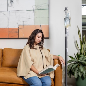 Puede incluir: Una mujer con una capa beige y pantalones vaqueros azules se sienta en un sofá de cuero marrón leyendo un libro. Una bolsa IV transparente cuelga de un soporte metálico junto al sofá. Una planta verde en maceta está en el fondo.
