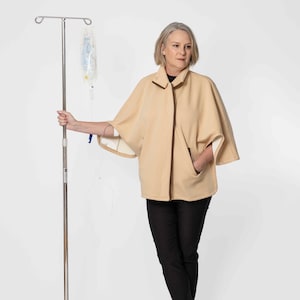 May include: A beige cape with brown trim and pockets. The cape is worn over a black top and black pants. The person is holding an IV stand with an IV bag.