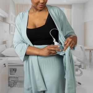 Puede incluir: Una mujer lleva una bata azul claro con un top negro debajo. Está sosteniendo una bolsa médica transparente en su mano izquierda. La bolsa está conectada a un tubo que va a su pecho.