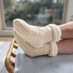 May include: Cream-colored cable knit slipper socks with a soft, white faux fur lining. The socks are cuffed and feature a textured cable knit pattern. The socks are resting on a light blue blanket.