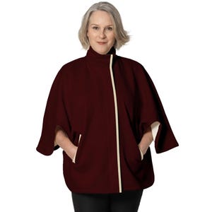 May include: A deep burgundy cape with a high collar and a contrasting cream-colored trim along the front opening and pocket edges. The garment features a loose, open-arm design with visible side pockets and a smooth, solid-colored fabric finish.