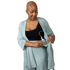 Puede incluir: Una mujer con una bata verde azulado claro y una camiseta negra, usando un sacaleches. La bomba está conectada a una botella transparente. La bata está abierta, mostrando la bomba y el pecho de la mujer. El fondo es blanco.