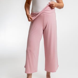 Puede incluir: Pantalones culotte rosa pálido con una cintura ancha. Los pantalones tienen un ajuste holgado y fluido y una longitud recortada, que termina justo debajo de la pantorrilla. La tela parece suave y ligera. Los pantalones se muestran sobre un fondo blanco.