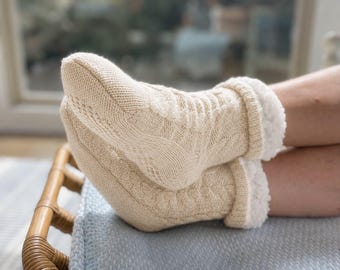 May include: Cream-coloured knitted slipper socks with a cable knit design and a soft, white faux fur lining. The socks are worn on a person's feet, resting on a light blue blanket. The socks appear warm and cosy.