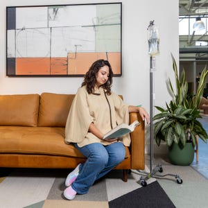 May include: A woman wearing a beige cape and blue jeans sits on a brown leather couch and reads a book. A medical IV pole with a clear bag is in the background.