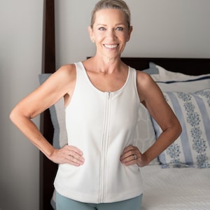 May include: A woman wearing a white, sleeveless zip-up top and light blue pants. The top has a high neckline and a full-length zipper. The woman is smiling and has her hands on her hips. The background includes a bed with blue and white pillows.
