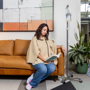 May include: A woman wearing a beige cape sits on a brown leather sofa and reads a book. A medical IV stand with a clear bag of fluid is in the background.