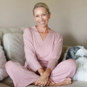 Puede incluir: Un conjunto de pijama rosa claro con una parte superior tipo envolvente y pantalones a juego. La parte superior de manga larga tiene cuello en V y los pantalones son holgados. La mujer está sentada con las piernas cruzadas en un sofá con cojines.