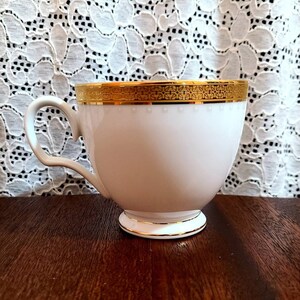 May include: A white teacup with a gold rim and a delicate floral pattern. The cup has a handle and a rounded shape.