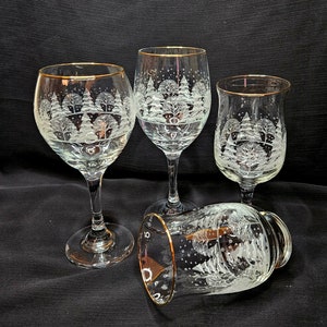May include: Four clear glass wine glasses with gold rims. Each glass has a white etched design of a winter scene with trees and snow.