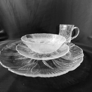 May include: A set of clear glass dishes with a floral pattern. The set includes a large plate, a smaller plate, a bowl, and a mug.