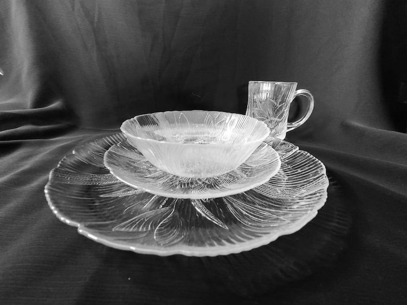 May include: A set of clear glass dishes with a floral pattern. The set includes a large plate, a smaller plate, a bowl, and a mug.
