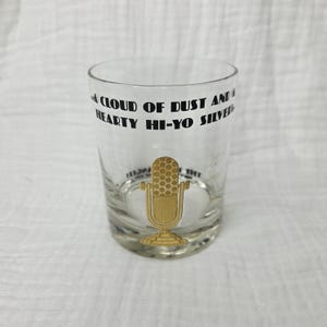 May include: A clear glass tumbler featuring a gold-colored vintage microphone graphic on the front. The text printed in black ink reads: A CLOUD OF DUST AND A HEARTY HI-YO SILVER. The glass has a thick base and a smooth, cylindrical shape.