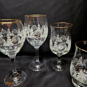 May include: Four etched glass wine glasses with gold rims. Each glass features a winter scene with trees and snow.