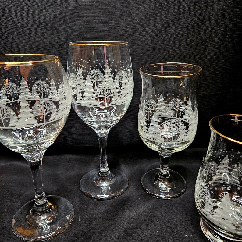 May include: Four etched glass wine glasses with gold rims. Each glass features a winter scene with trees and snow.