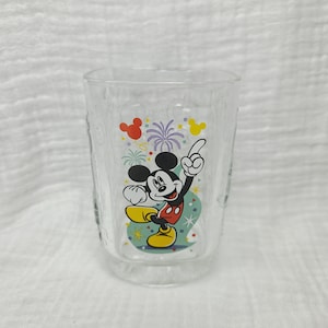 May include: A clear glass tumbler featuring a colorful graphic of Mickey Mouse pointing upward. The design includes red and yellow balloons, purple fireworks, and small confetti shapes surrounding the character on a light teal background.