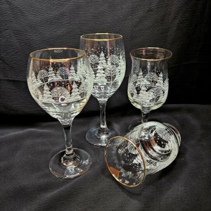 May include: Three clear glass wine glasses with gold rims and etched winter forest designs. The glasses are on a black surface.