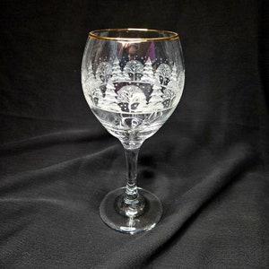 May include: A clear glass wine glass with a gold rim and a frosted design of a winter scene with trees.
