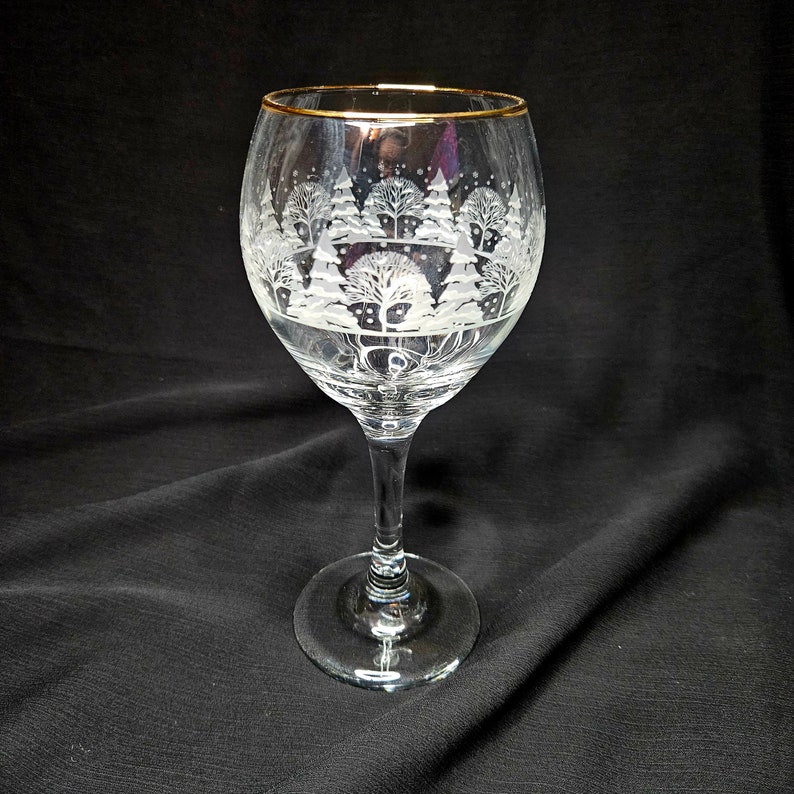 May include: A clear glass wine glass with a gold rim and a frosted design of a winter scene with trees.
