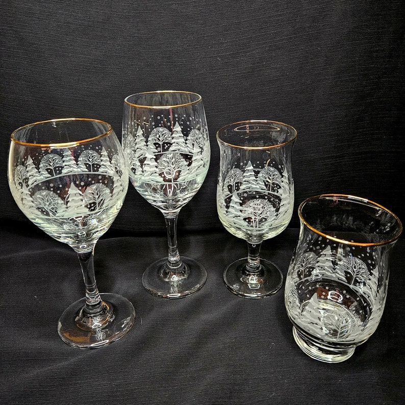 May include: Four etched glass wine glasses with gold rims. Each glass features a winter scene with trees and snow.