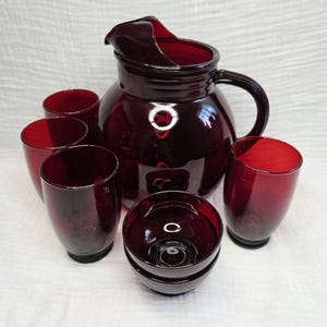 May include: A set of deep red glass drinkware consisting of a large pitcher with a curved handle and pouring spout, four tall drinking glasses, and two small bowls. The glassware features a smooth, translucent finish in a consistent ruby hue.